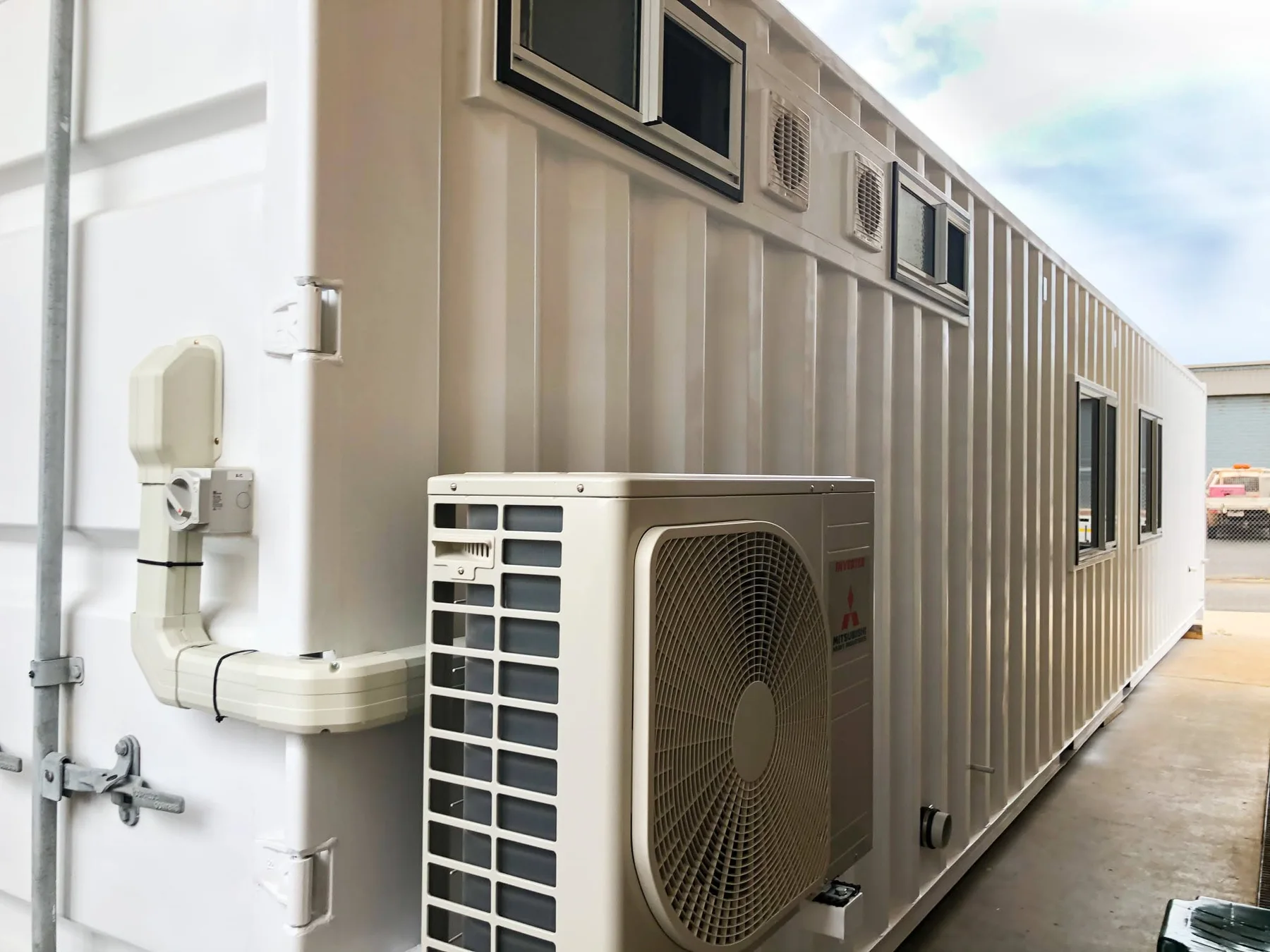 Outdoor split-system air-conditioning unit mounted beside windows and ventilation grilles on a converted container