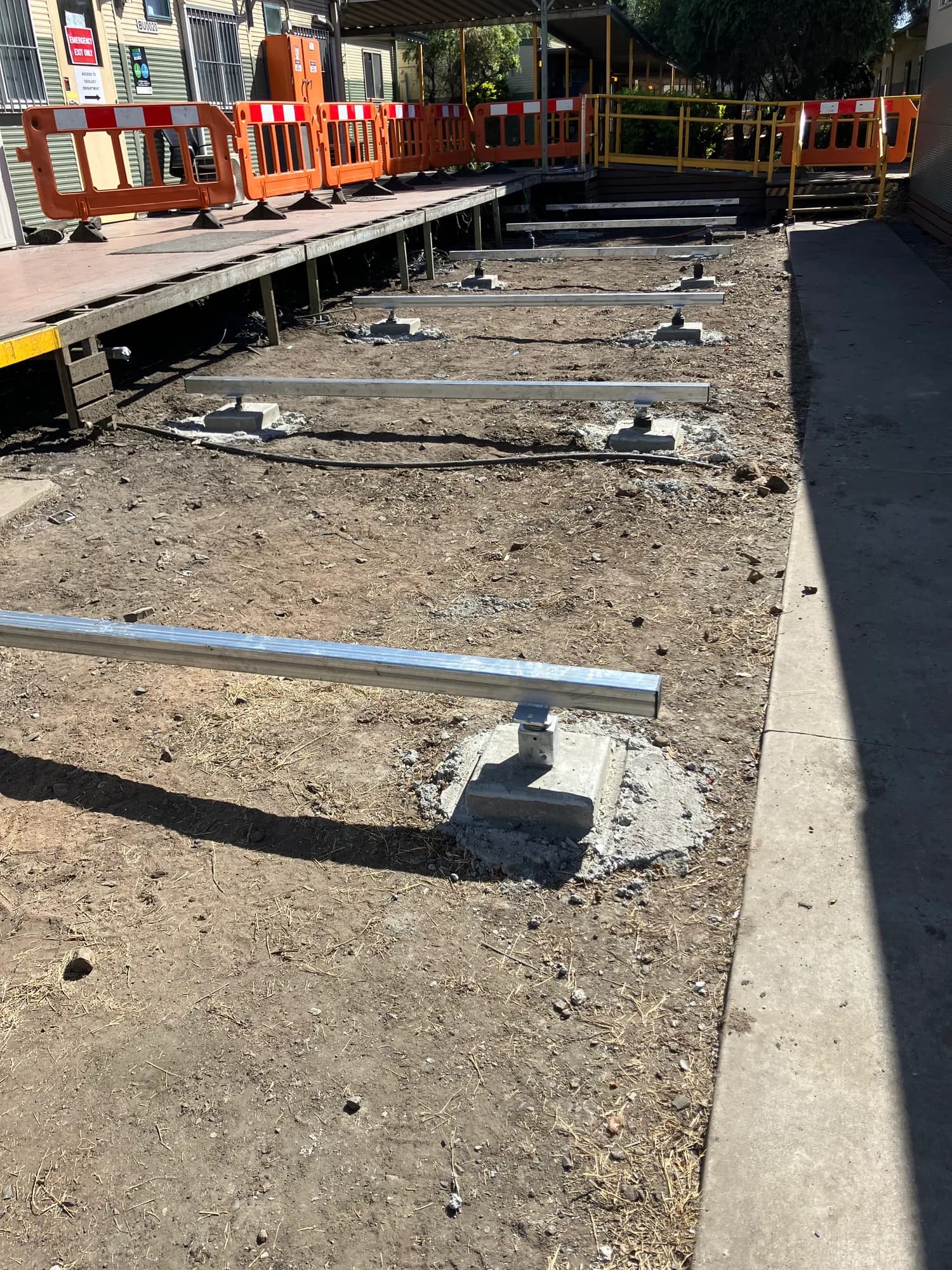 Concrete footing pads and steel building supports in a barricaded installation area beside existing decks