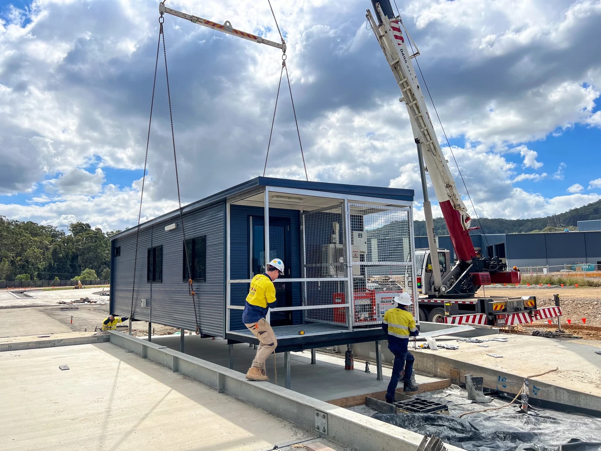 Crane setting a navy transportable building onto steel stumps while two installers guide it into place