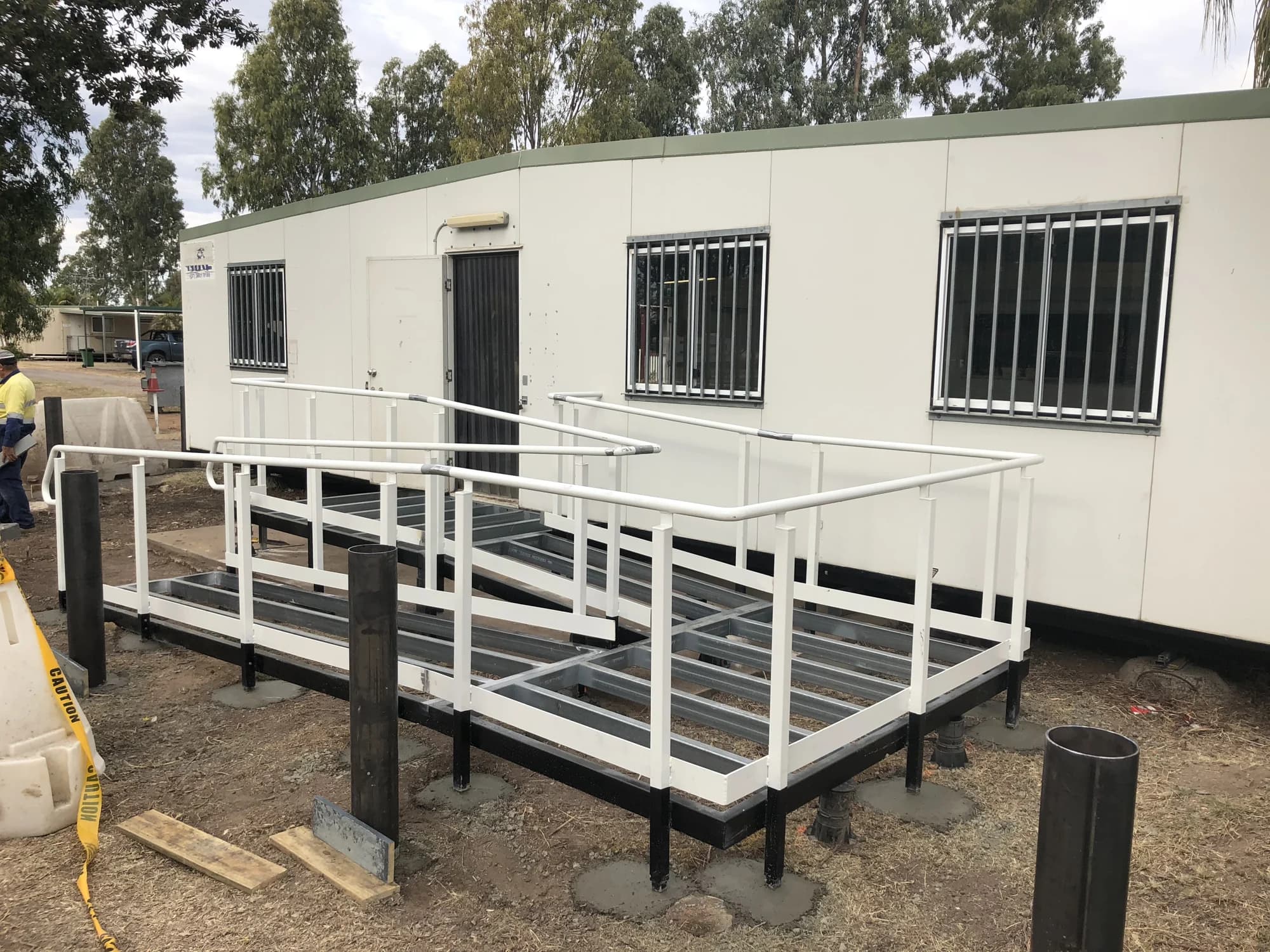 Ramp under construction, with exposed steel floor framing and white handrails beside an existing building