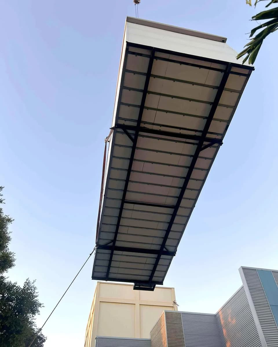 Underside of a portable building suspended from crane rigging above existing buildings