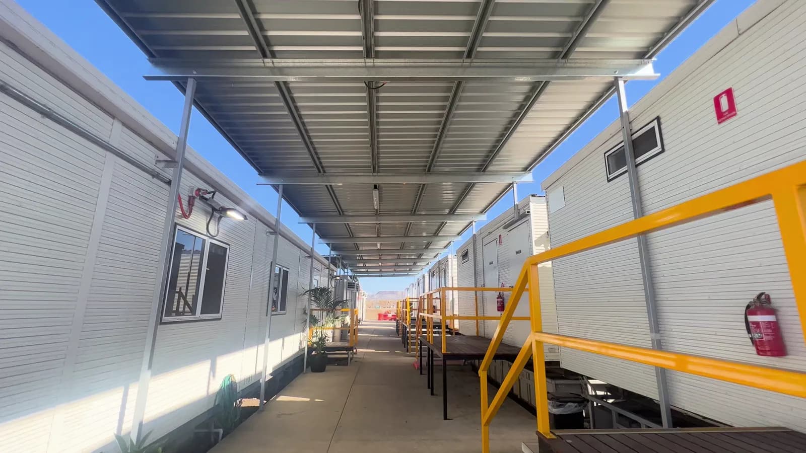 Concrete corridor beneath steel awnings with yellow-railed stairs and landings beside modular buildings